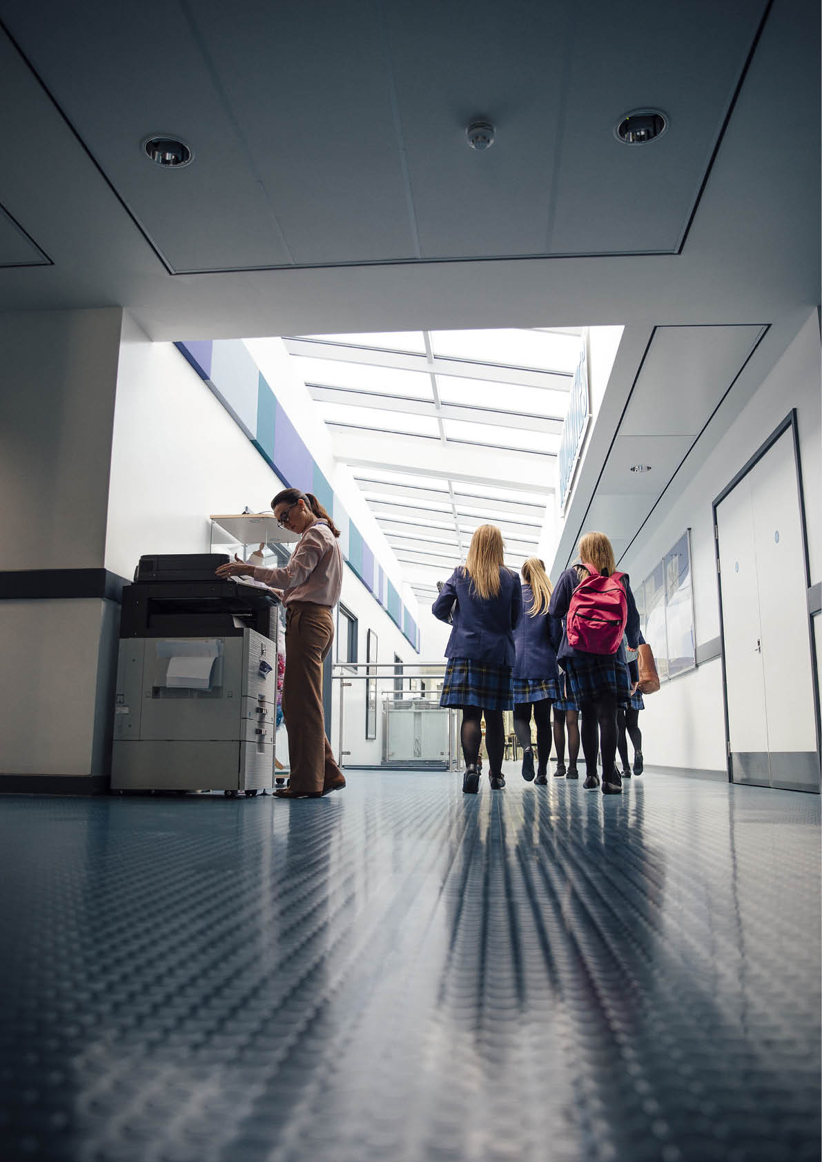 Group of students are walking down the corridor of their school. There is a female teacher using the printer.