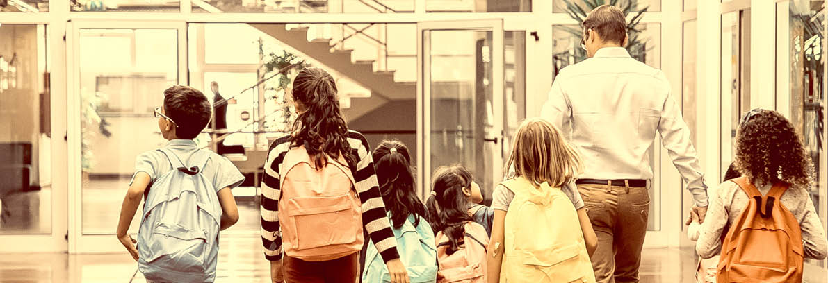 Schoolkids with colorful backpacks and male teacher walking through school hallway. Back view, full length. Education or back to school concept