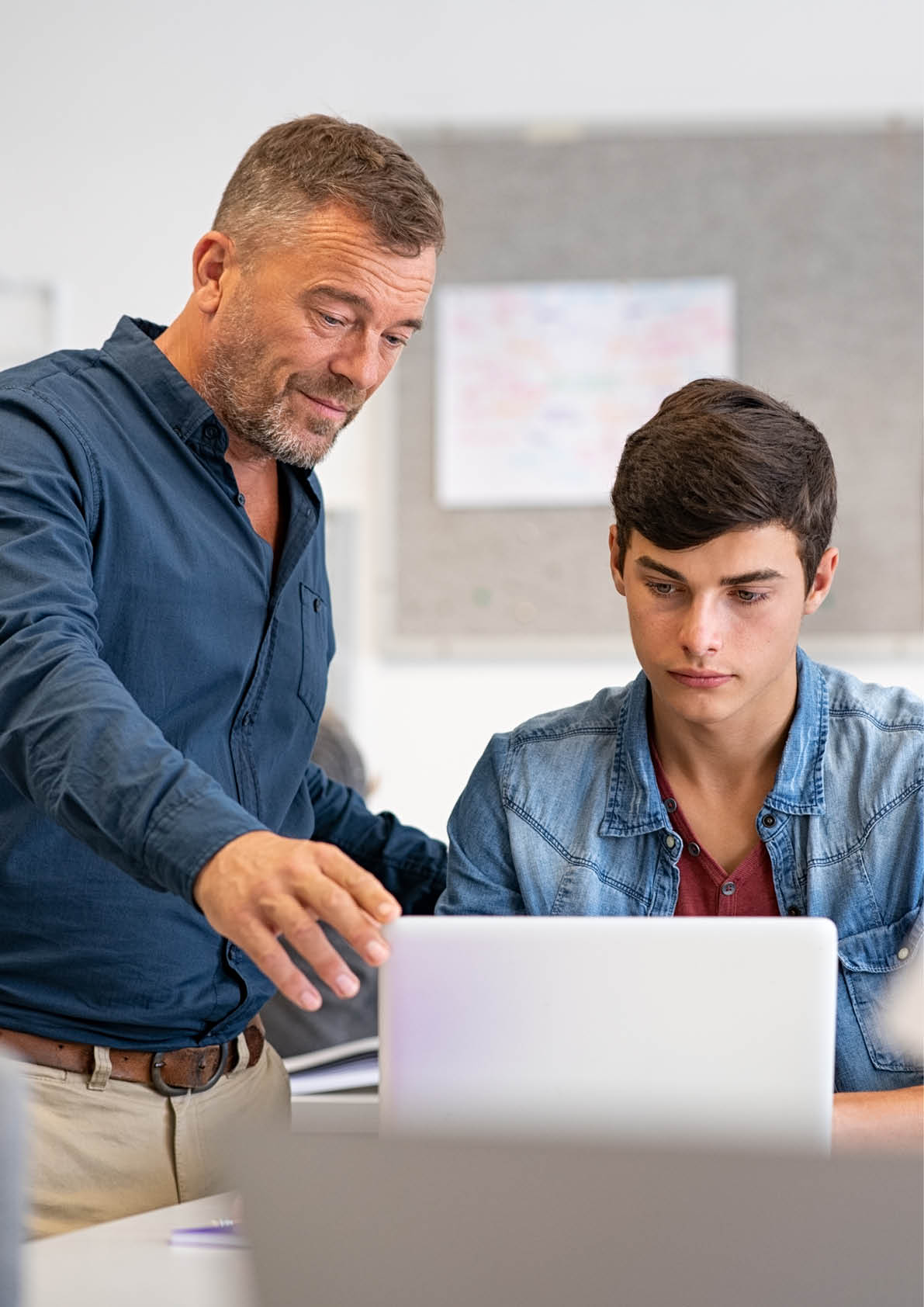 Professor assisting college student with laptop in classroom during computer lesson. Teacher talking and explaining something to guy sitting at desk. Mature man lecturer helping high school young man with laptop during lecture in university.