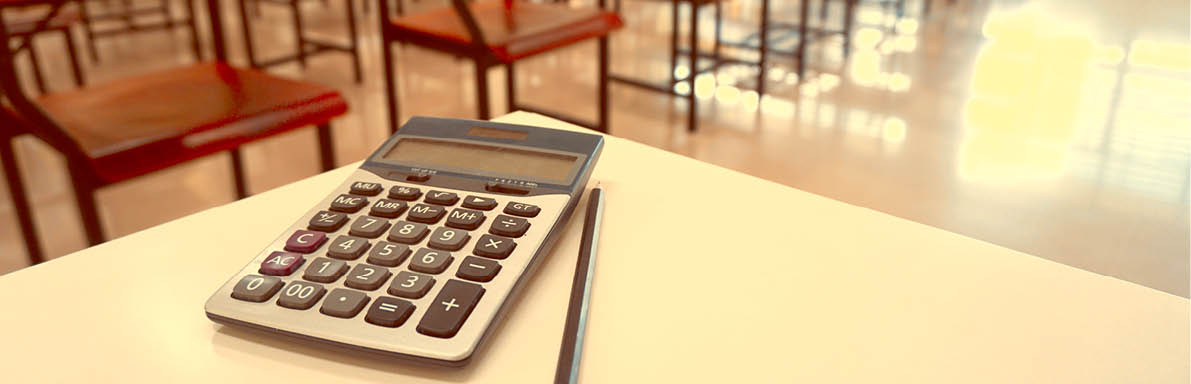 The calculator and pencil on the teacher's desk with the wood lecture chairs place distance in the empty classroom. Back to school, business, and finance accounting concept. Copy space in Thailand.
